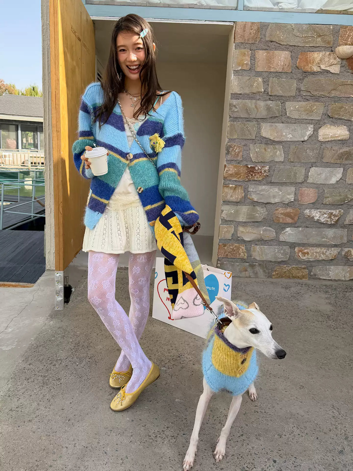 Woman in the blue and navy striped cardigan holding a paper cup, a whippet in a blue and yellow striped knit beside her