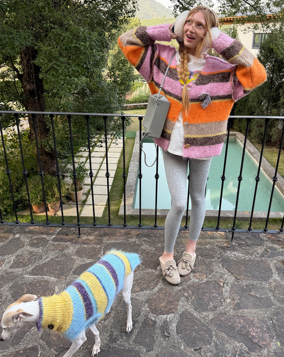 Woman lifting her braids in the pink and orange striped cardigan, a whippet in a blue and yellow striped knit on the stone terrace