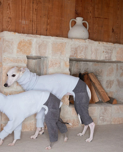 Rear three-quarter view of two whippets in pale blue fleece tops and heather grey cuffed hind legs, showing the hip pocket patch and the open cut around the back leg, beside a stone fireplace stacked with logs